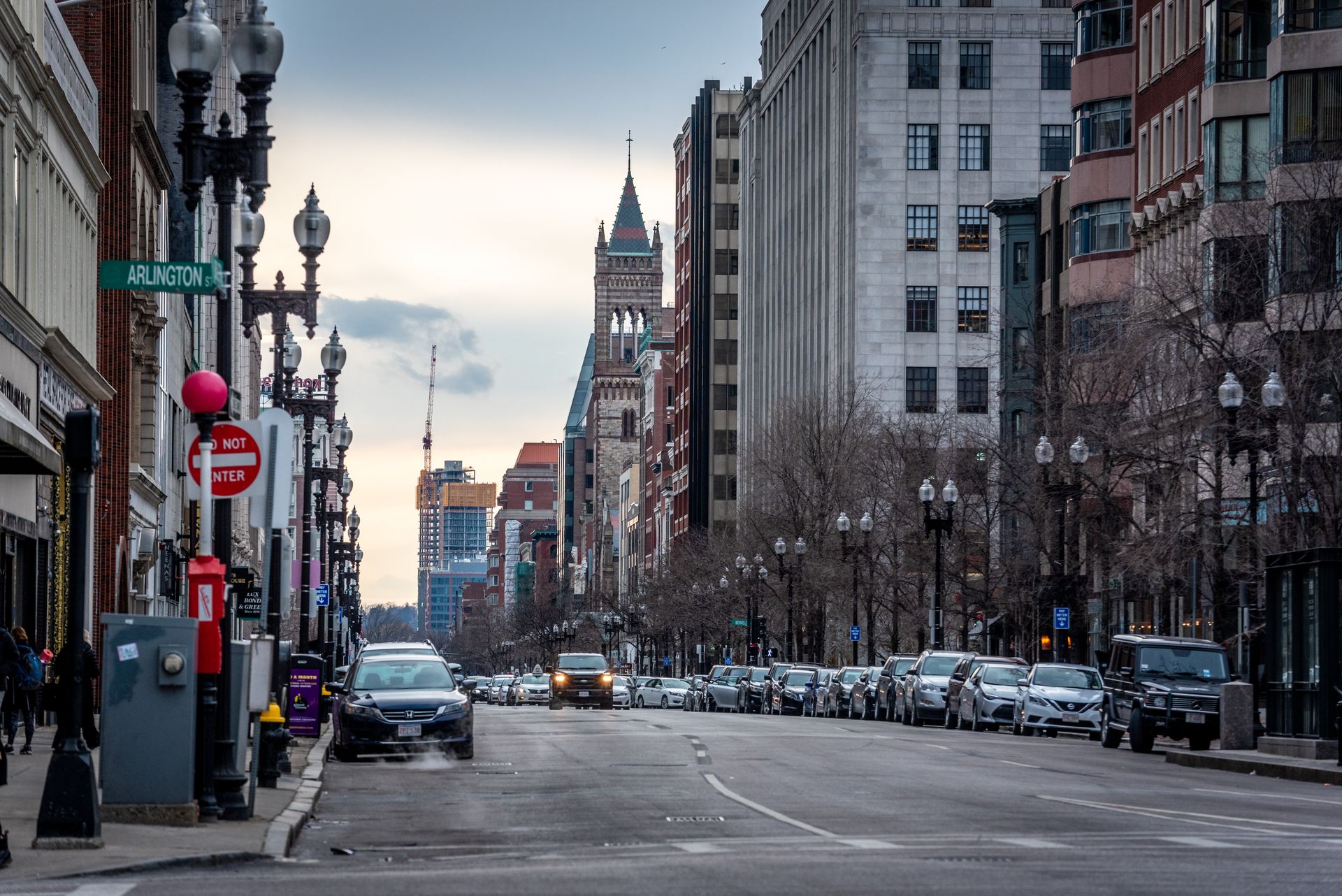 City street with parked cars, ornate street lamps, and a stone church tower in the distance